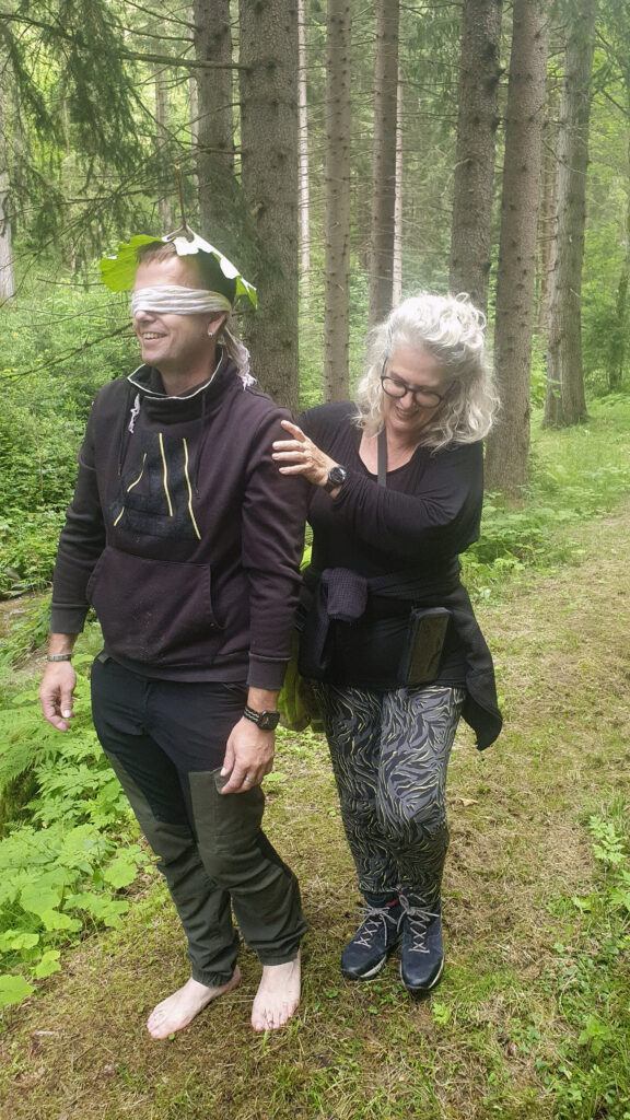 Mann, barfuß wird mit verbundenen Augen von einer Frau durch den Wald geführt.
