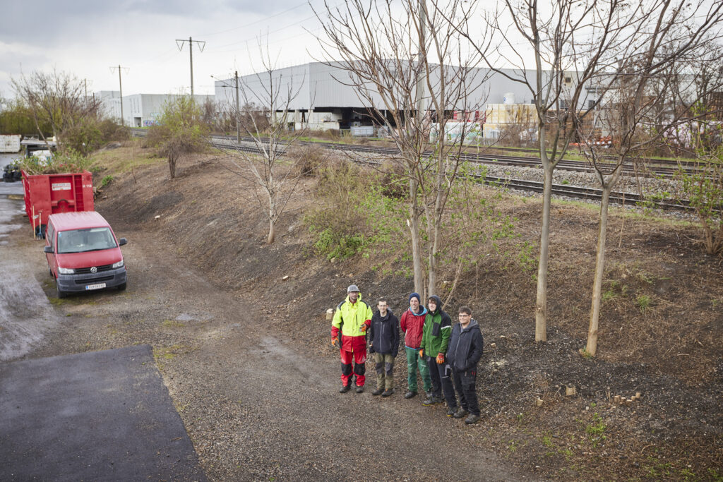5 Personen, draußen in Arbeitskleidung stehen vor freigeschnittenen Bäumen im Gewerbegebiet.