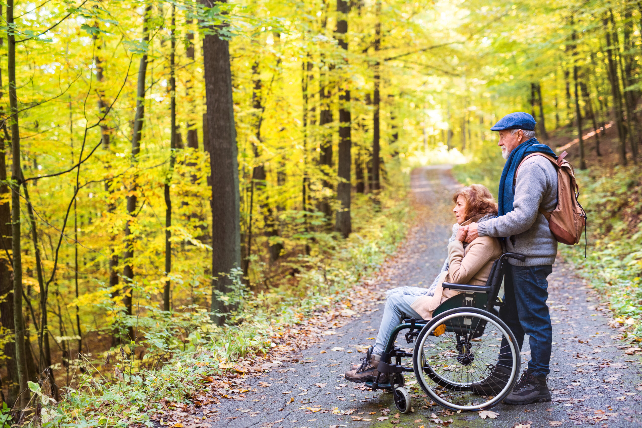 Seitliche Ansicht einer Frau im Rollstuhl sitzend, hinter ihr ein Mann stehend mit den Händen auf ihren Schultern. Sie stehen quer auf einem asphaltierten Weg im Herbstwald.