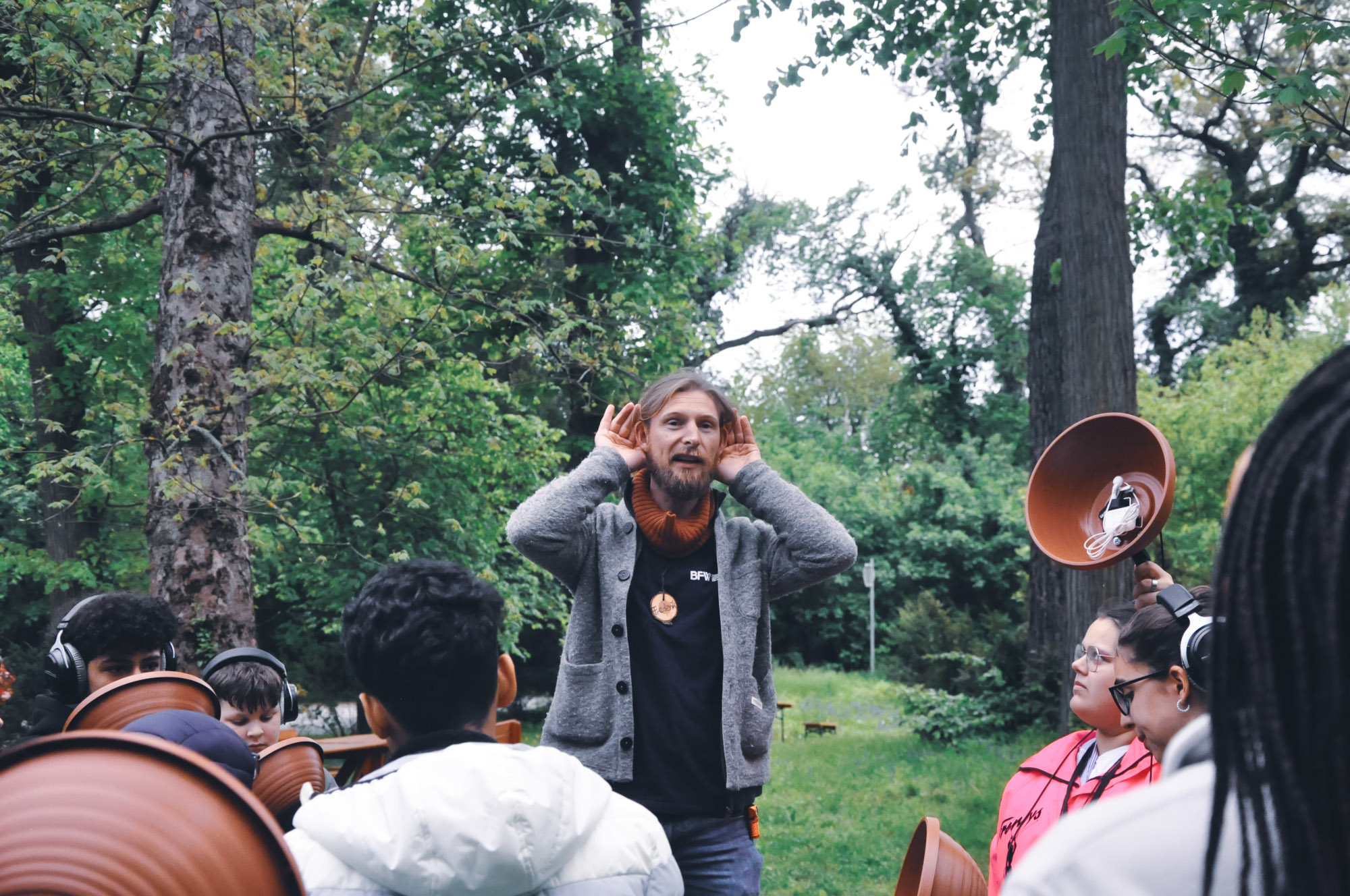 Frederick Sachser steht mit den Schüler:innen in der Natur und legt beide Hände an die Ohren. Die Schüler:innen halten ihre Plant-Pot-Mics in der Hand.