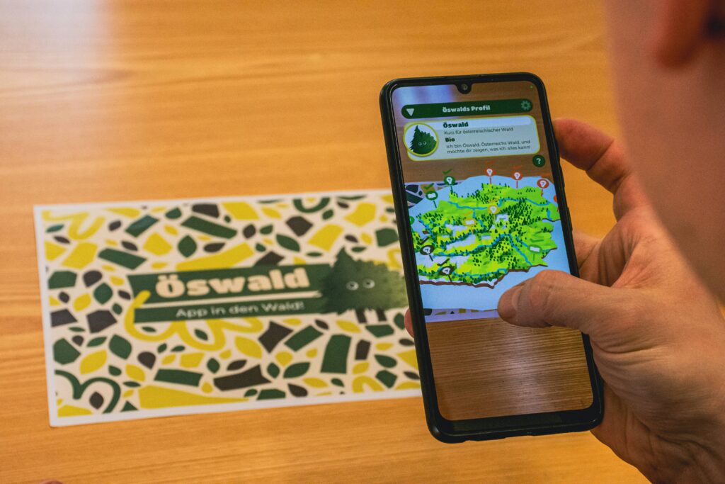 Handy-Bildschirm auf dem eine Österreichkarte zu sehen ist. Im Hintergrund ein Bild mit dem Wort Öswald, dass gesannt wird.