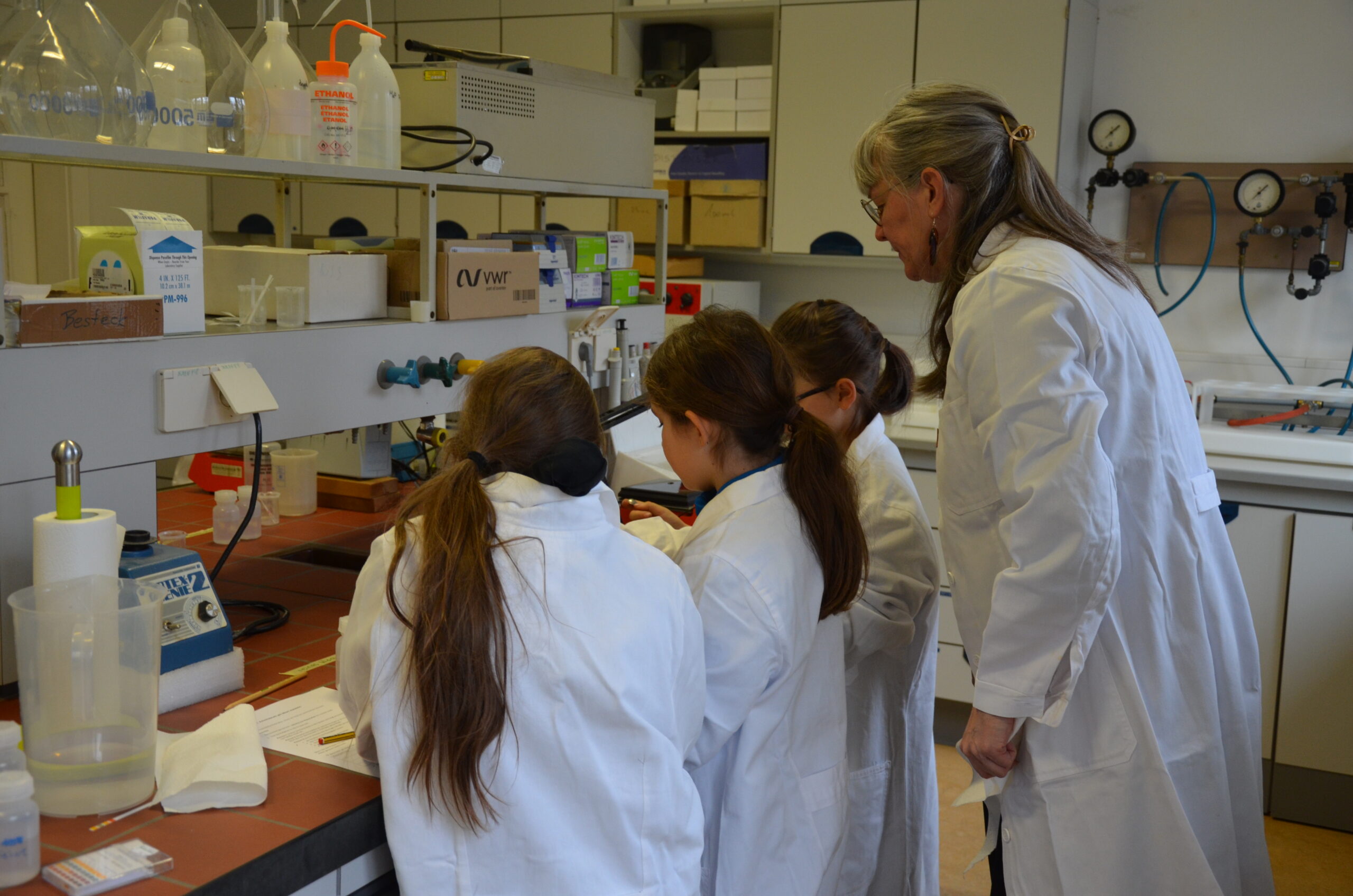 Eine Erwachsene steht hinter drei Mädchen in einem Labor und beobachtet ihr Tun.