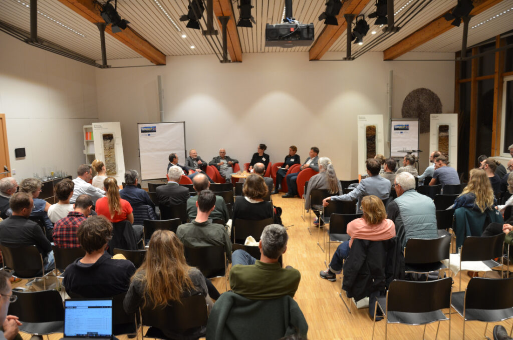 Mehrere Leuten sitzen bei einer Tagung in einem Saal. Vorne gibt es eine Sesselreihe mit Diskussionsteilnehmern