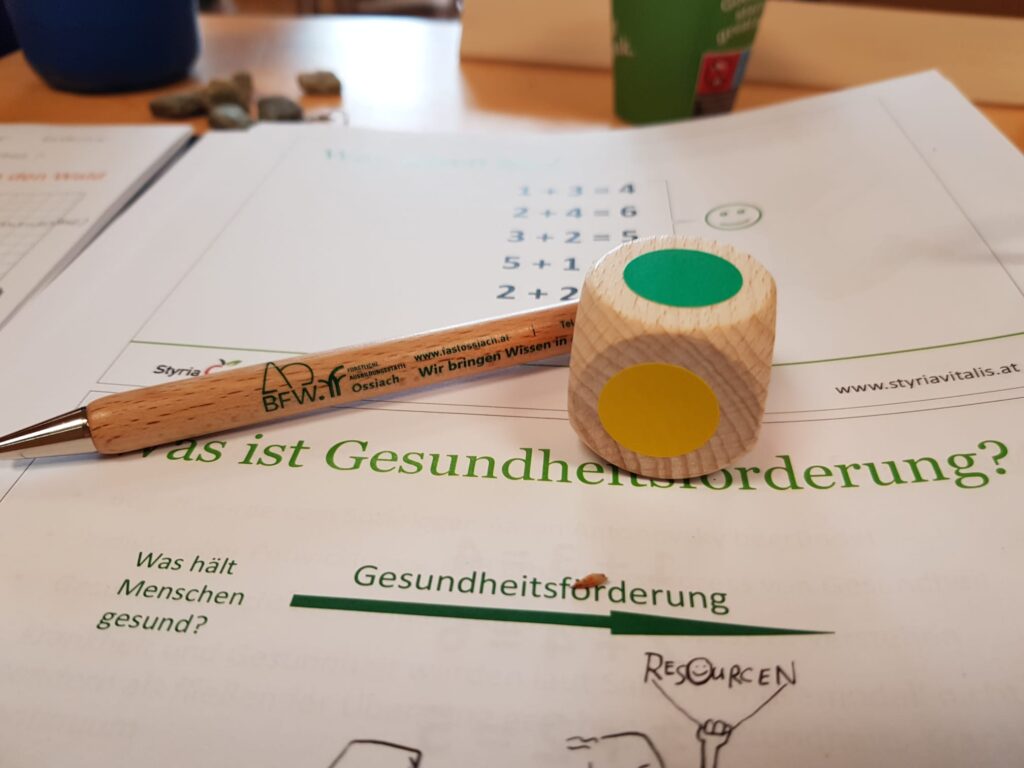 Skrift mit Titel "Was ist Gesundheitsförderung?", Würfel zeigt oben grüne Farbe, vorne gelb, Holzkugelschreiber mit Aufschrift BFW Ossiach