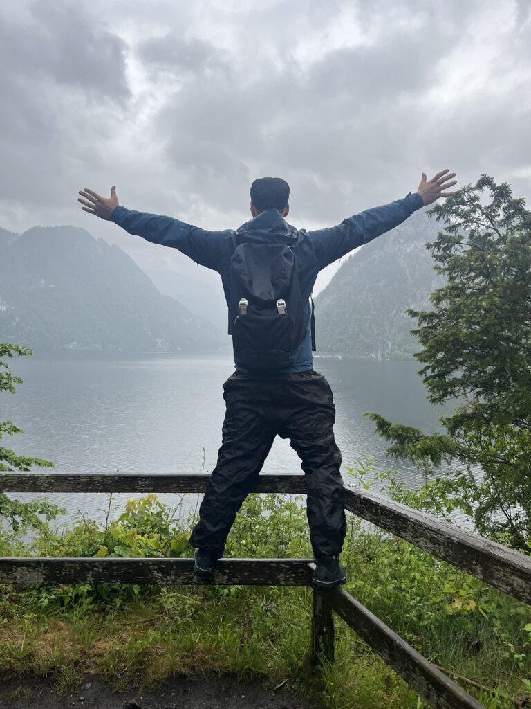 Mann steht auf einer Abgrenzung, ausgebreitete Arme und Blick auf einen See und wolkenverhangene Berge