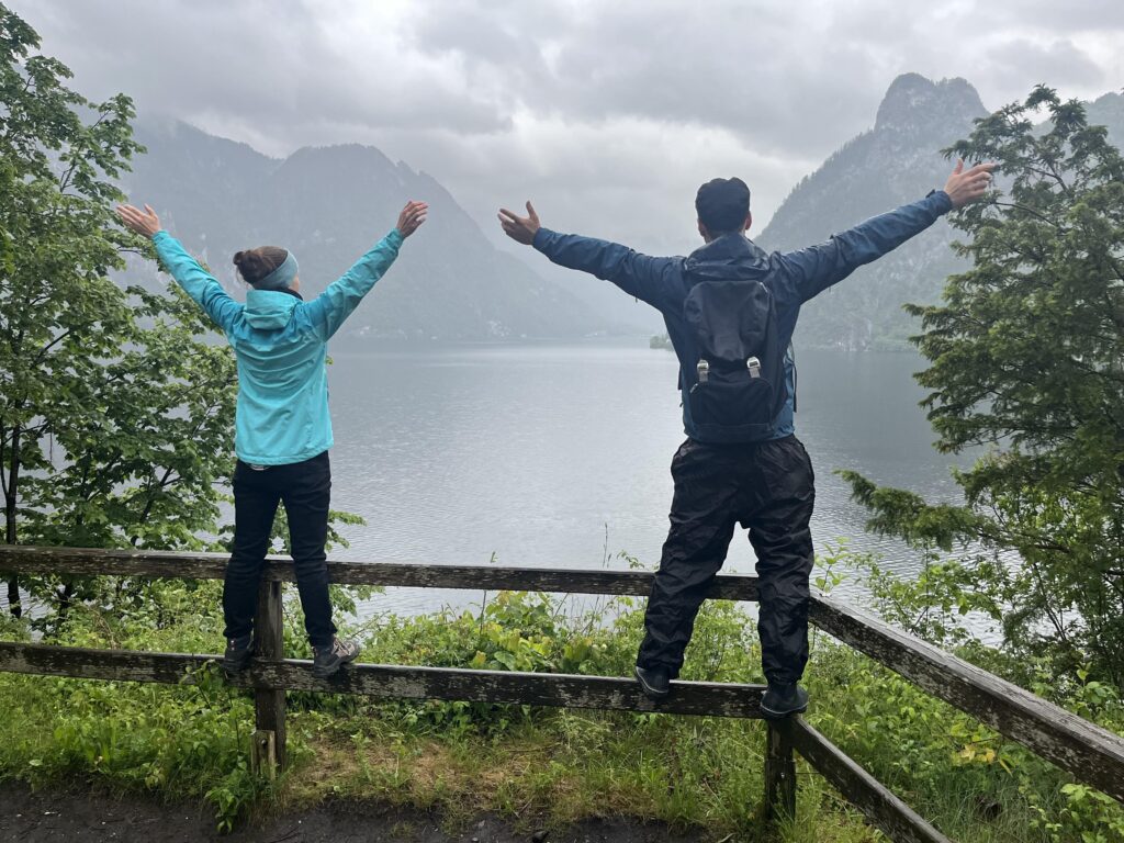 1 Frau und 1 Mann stehen outdoor auf einem Geländer, mit Rücken zu uns, Arme ausgebreitet, und blicken auf den See und die wolkenverhangenen Berge