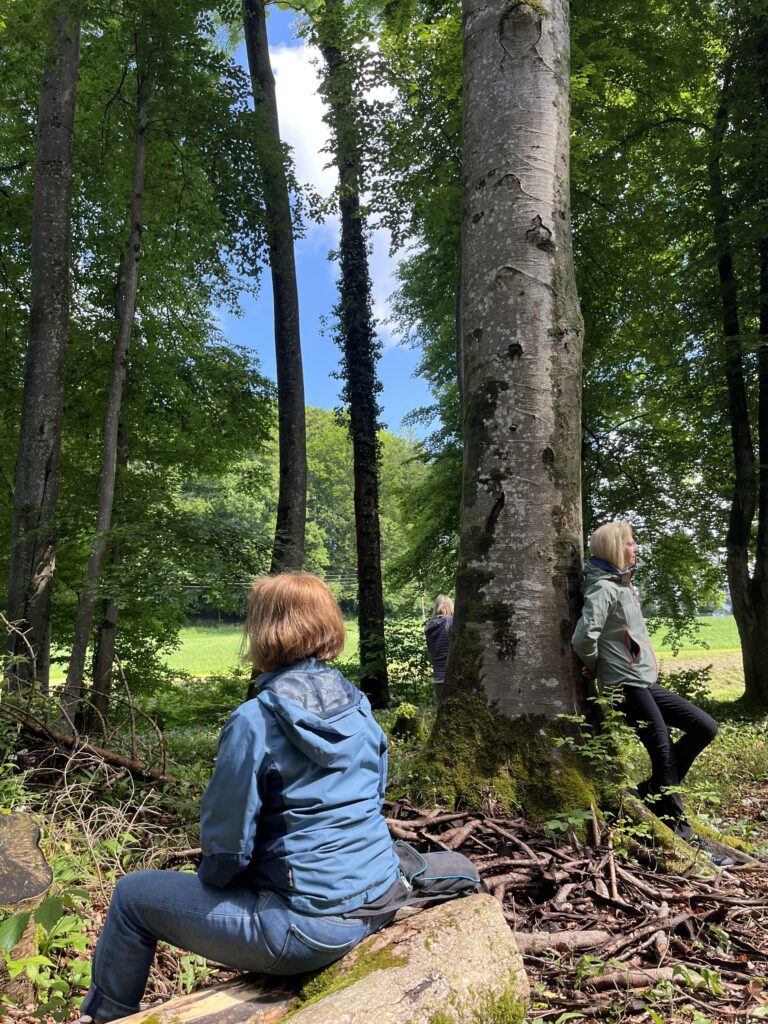 Personen im Wald, eine sitzt im Vordergrund, eine steht an einen Baum gelehnt.