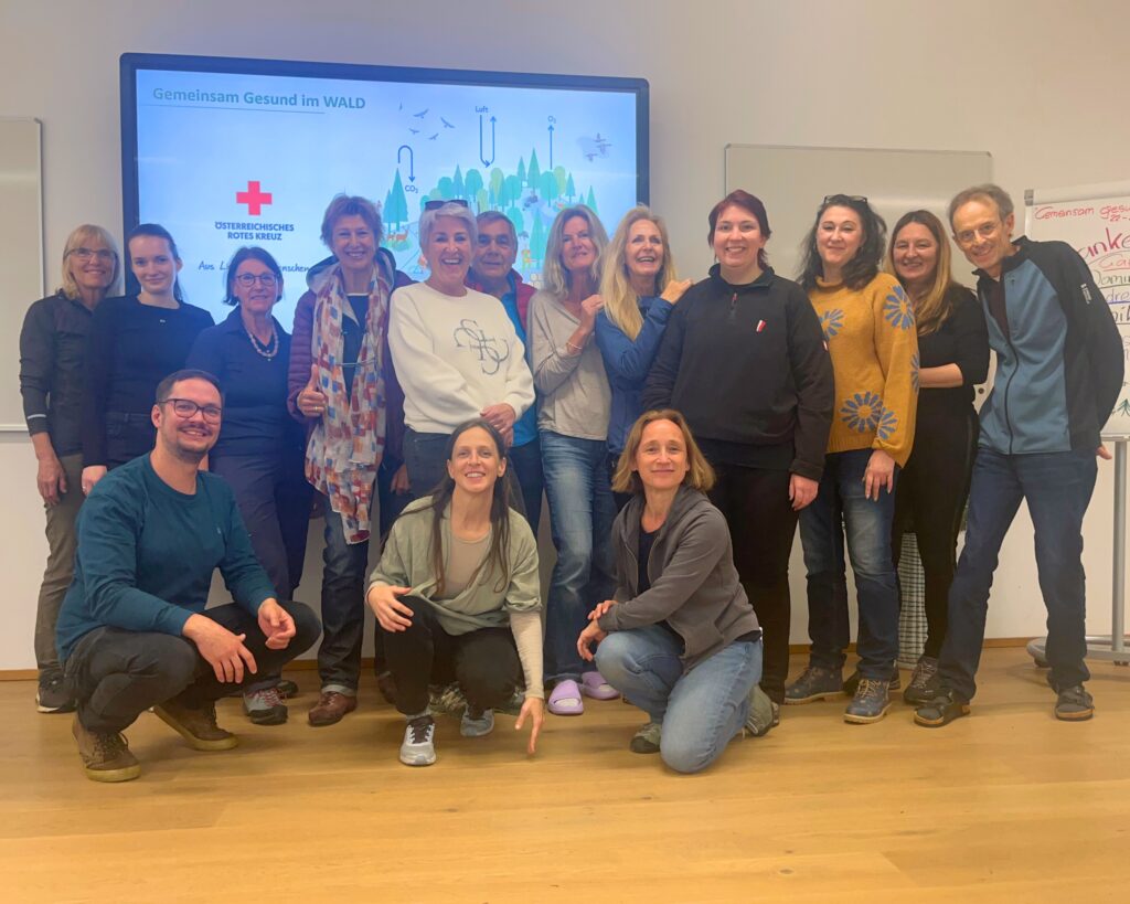 Gruppe von Frauen und Männern im Innenraum. Im Hintergrund zu lesen "Gemeinsam Gesund im Wald" und Logo Rotes Kreuz.
