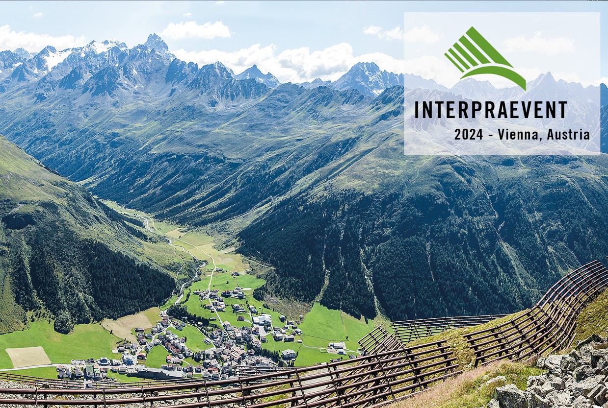 Blick auf ein Dorf im Tal, An den Hängen Schutzwald und ein Lawinenschutzzaun. Im rechten oberen Eck das Logo von Interpraevent 2024