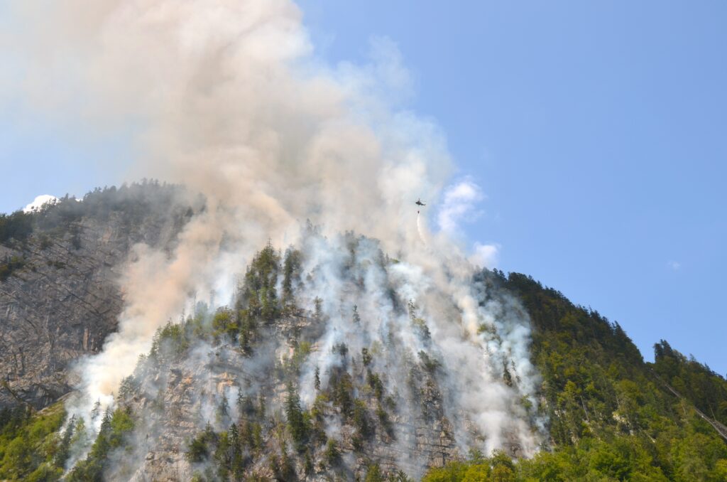 Brennender Schutzwald auf einem Berghang. Ein Löschhubschrauber versucht den Brand einzudämmen